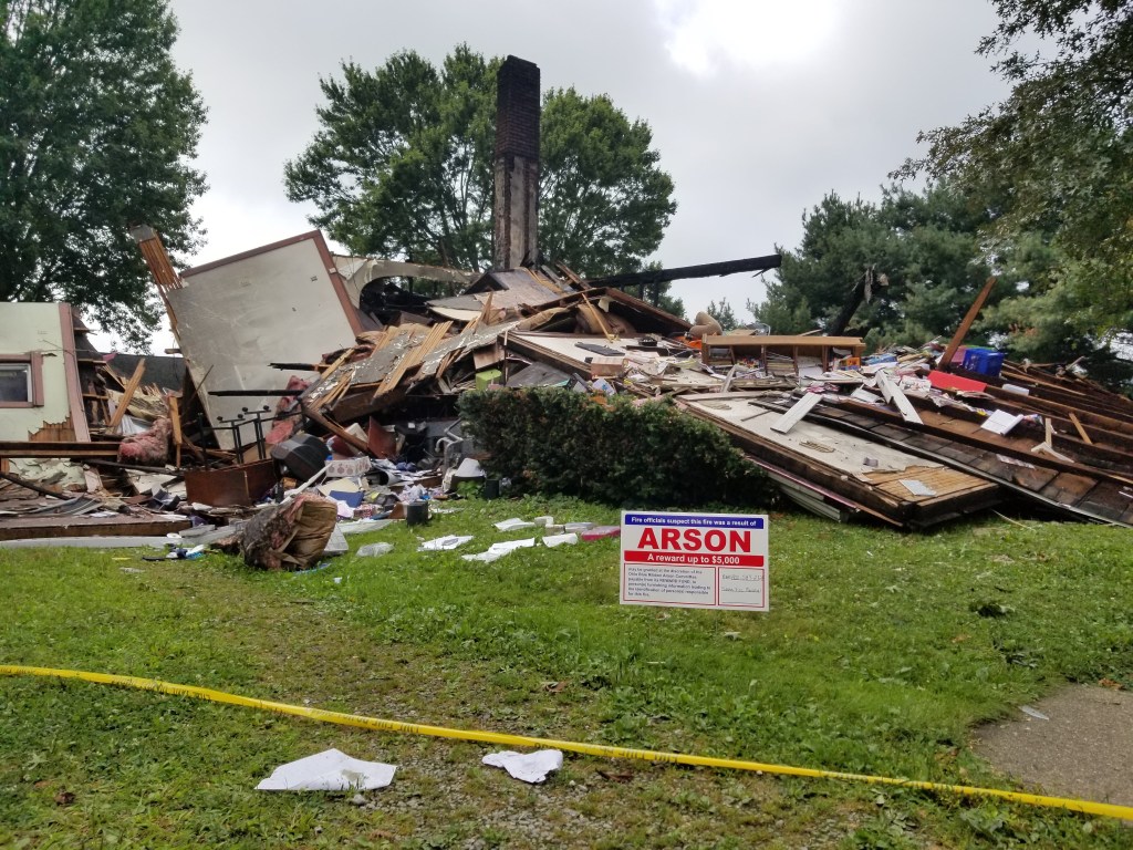 A sign reading "ARSON" stands in the lawn in front of the ruins of a home destroyed in an explosion