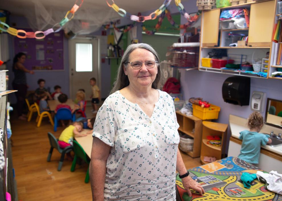 A child care center director, a woman with long gray hair, poses for a portrait at the facility, with children in the background